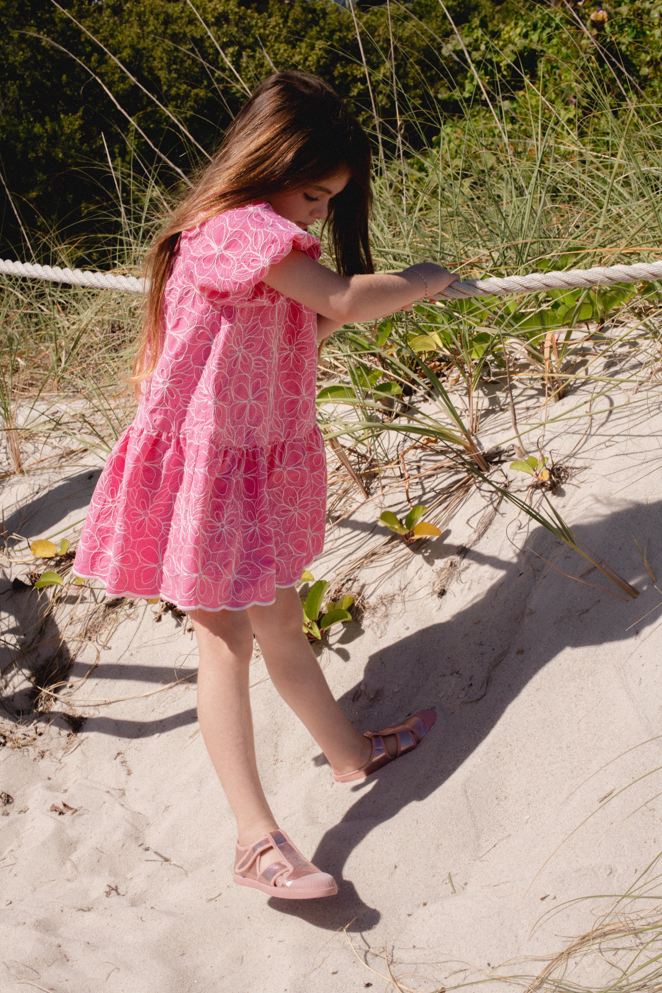 Young girl in a pink dress standing on a sandy beach with grass and rope in the background.