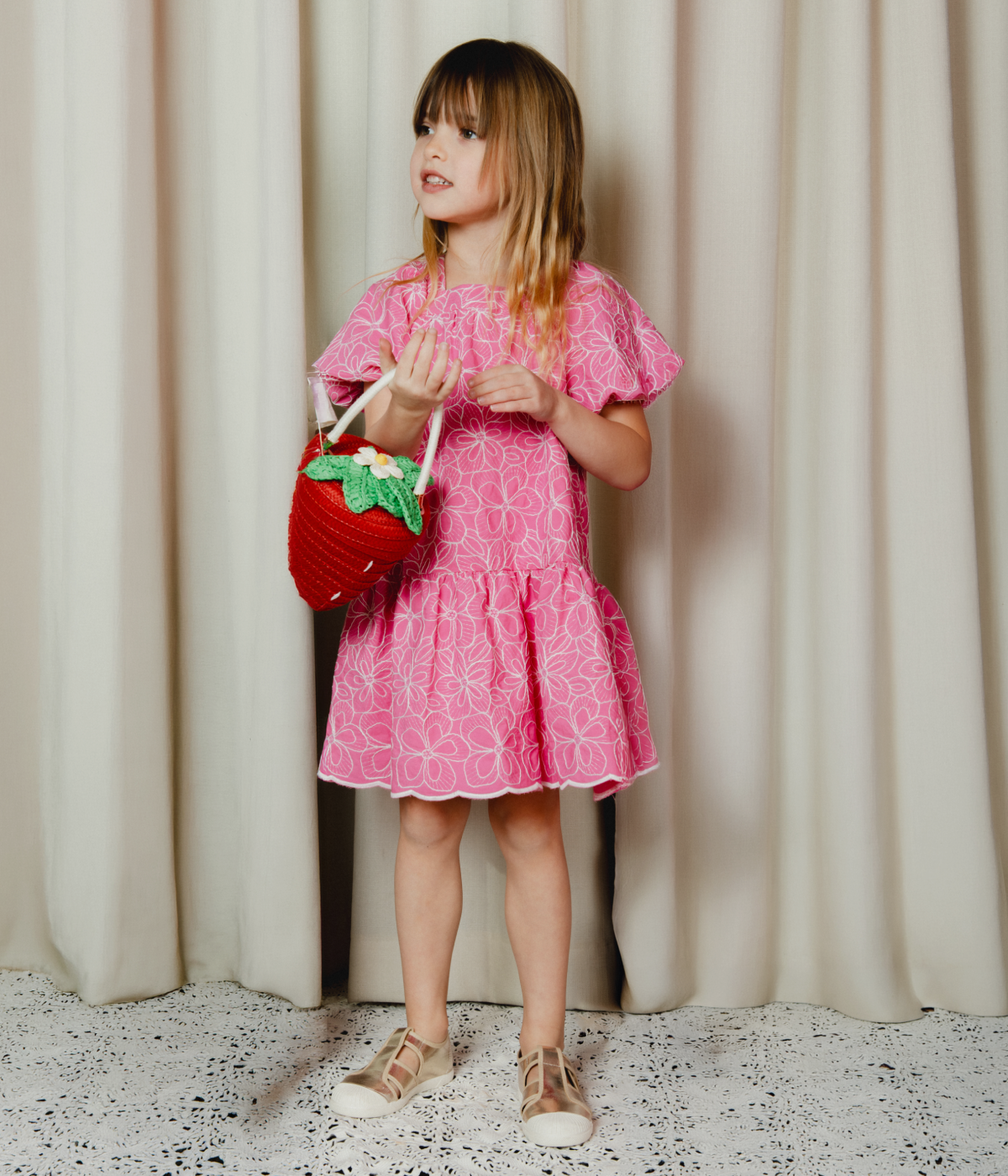 Young girl in a pink dress holding a red bag with a floral design against a white curtain background