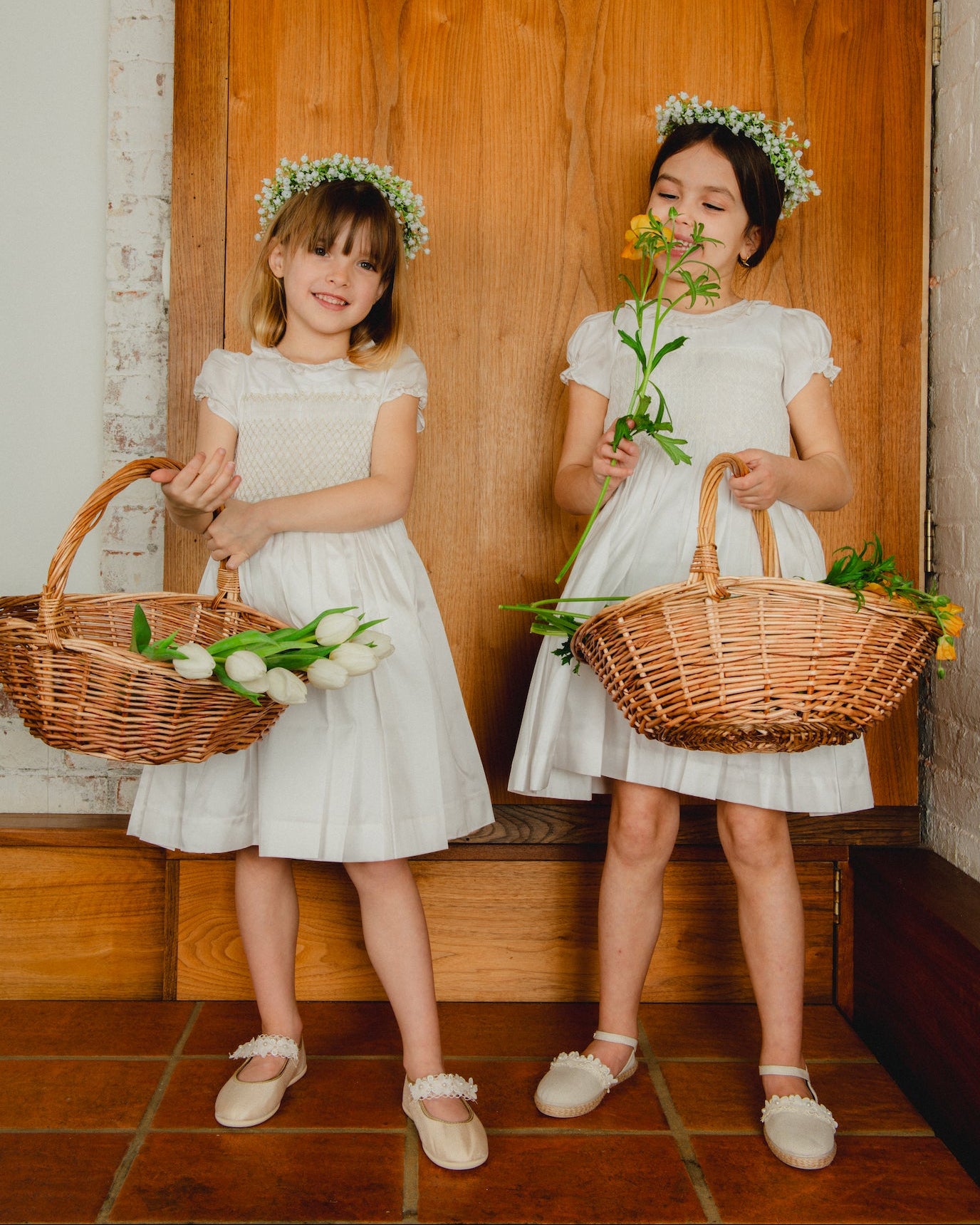 Two young girls in white dresses with flower crowns holding baskets and flowers against a wooden background.